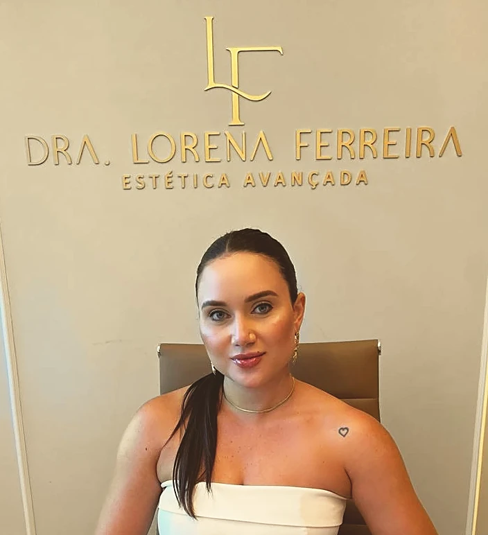 Dra. Lorena Ferreira sentada à mesa do consultório, sob o letreiro dourado com o próprio nome na parede