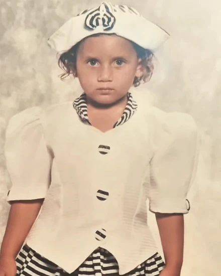 Lorena criança, de chapéu e casaquinho brancos com detalhes listrados, em foto de estúdio antiga
